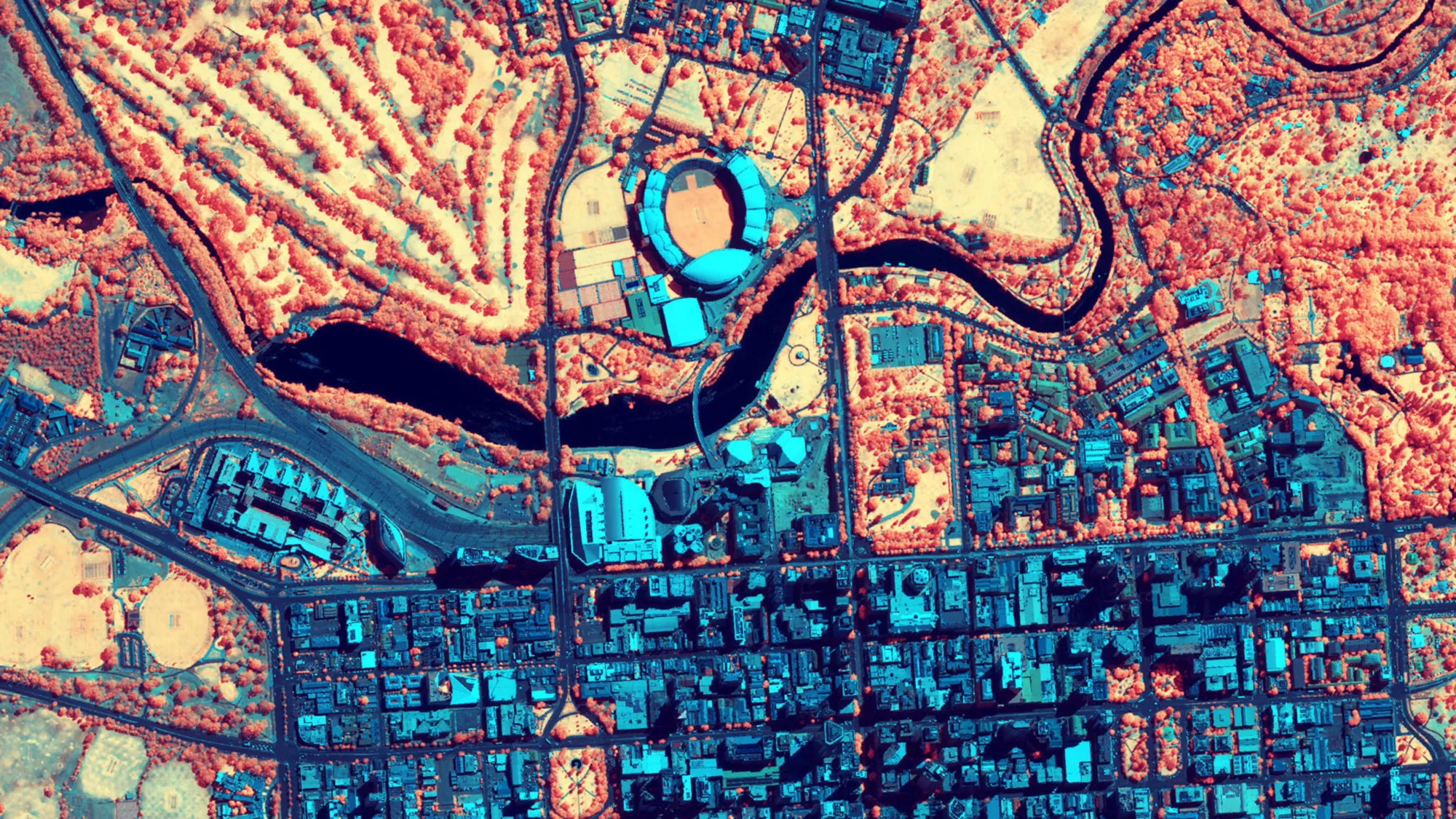 Color-coded satellite map of a city with blue, orange, and red hues, showing a river, green spaces, and a stadium-like structure in a grid-like urban layout.