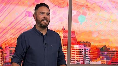 A young man in a blue button-down shirt against a pink and red abstract background of a building