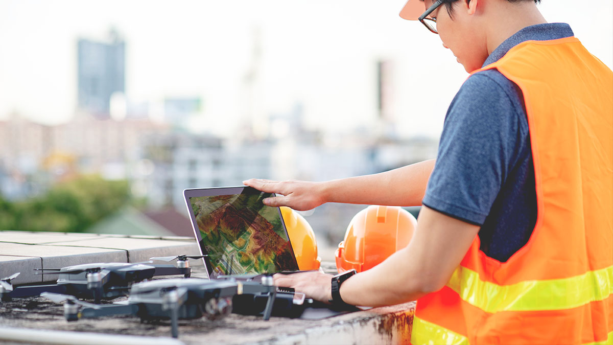 An on-site worker in a yellow safety vest connects a drone to a laptop