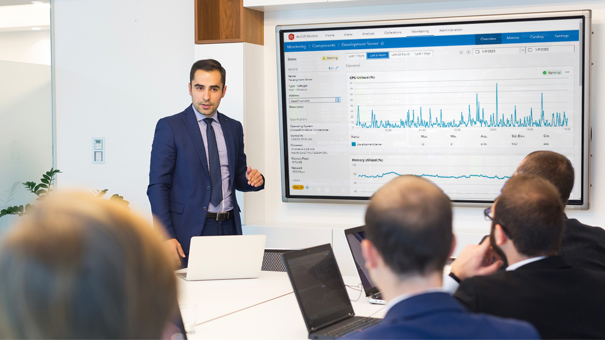 A group of men in a conference room with their laptops and another man standing next to a large monitor displaying a graph