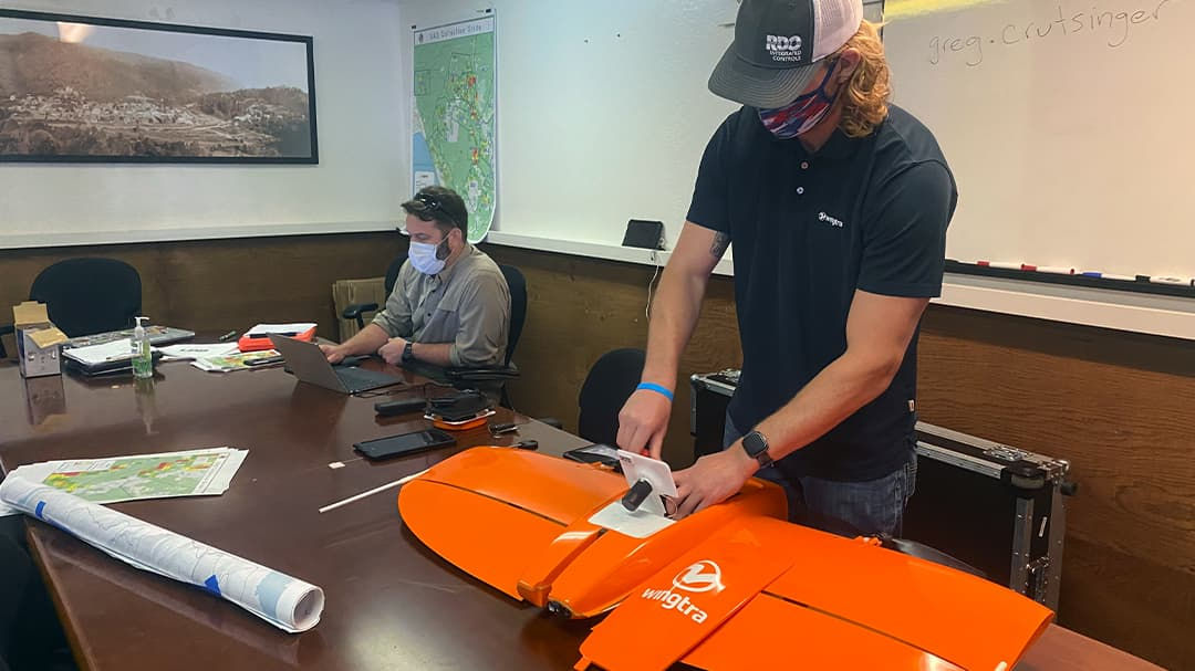 Two workers wearing face masks at a conference desk, one working on a laptop and the other working over a bright orange drone